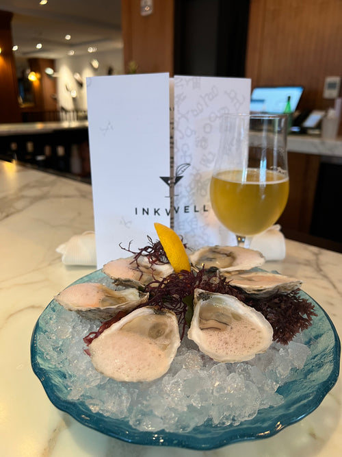 Plated dish of oysters on a bed of ice with a glass of white wine and 'INKWELL' menu card.
