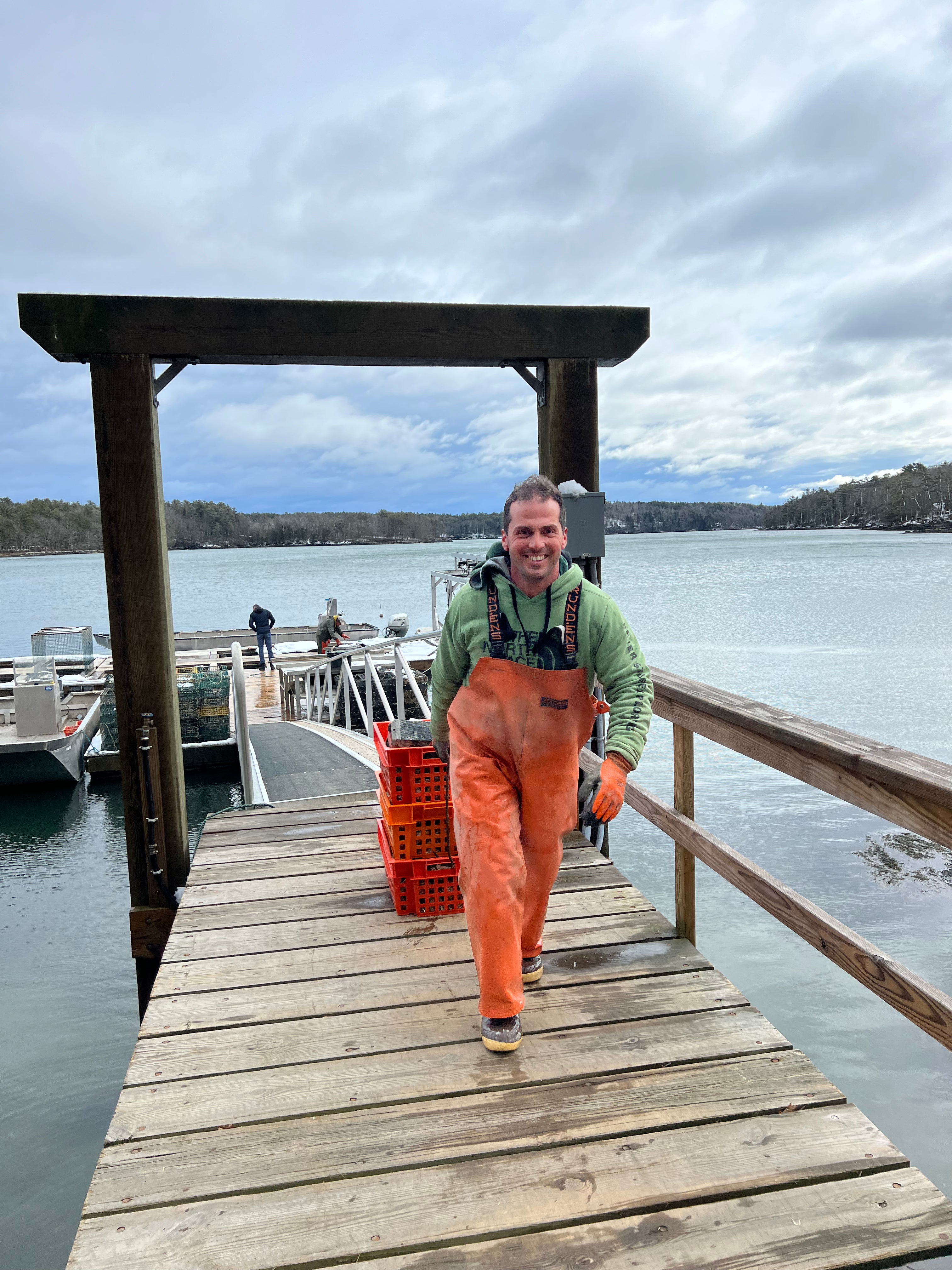 Person in orange overalls walking on a dock with a crate, against a cloudy sky.