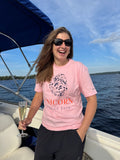 Person on a boat wearing a pink Unicorn Oyster t-shirt with a graphic design, holding a glass of champagne.